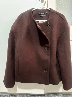 Brun ulljacka med stora knappar - Mörkbrun jacka i ull med rak passform och rund halsringning. Jackan har tre stora knappar framtill och diskreta fickor i sidorna. Enkel och stilren design som funkar till många olika looks.