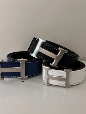 Svart, blå och vit skinnbälte med H-spänne - Tre stilrena bälten i svart, marinblått och vitt skinn med stora H-formade metallspännen. Perfekta för att lyfta vilken outfit som helst och ger en lyxig känsla. Snygga kontrastsömmar längs kanterna för extra detalj.