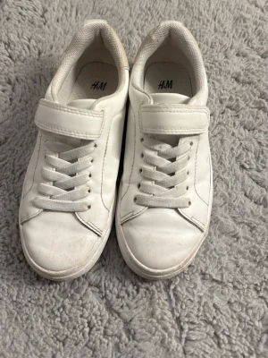 Vita sneakers med kardborre från H&M - Stilrena vita sneakers från H&M med klassisk låg profil. Skorna har både snörning och en praktisk kardborrestängning upptill. Tillverkade i syntetmaterial med vit sula och diskret design som passar till det mesta.