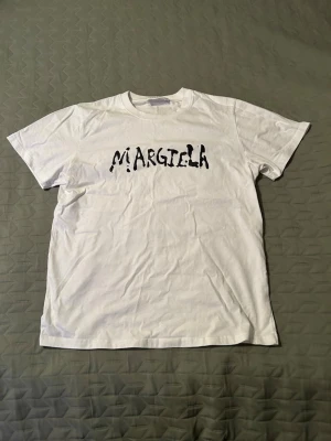 Vit Margiela t-shirt med tryck - Snygg vit t-shirt med svart Margiela-tryck framtill. Klassisk passform med rund hals och korta ärmar. Tillverkad i mjuk bomull som känns skön mot huden. Perfekt för dig som gillar minimalistisk och trendig stil.