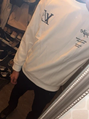 Vit sweatshirt med tryck - Säljer en vit sweatshirt med tryck på både bröstet och ryggen. Tröjan har en avslappnad passform och ribbade muddar vid ärmslut och nederkant. Materialet ser ut att vara mjukt och bekvämt, perfekt för en chill look. Inga synliga fickor eller huva.