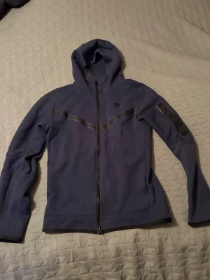 Mörkblå Nike hoodie med dragkedja - Snygg mörkblå Nike hoodie med hel dragkedja och huva. Jackan har svarta detaljer över bröstet och en ficka med dragkedja på ena ärmen. Tillverkad i mjukt material, perfekt för en avslappnad stil.