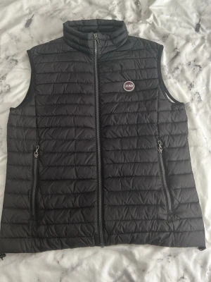 Svart dunväst från Colmar - Snygg svart dunväst från Colmar med quiltad design och hög krage. Västen har två dragkedjeförsedda fickor framtill och en diskret logga på bröstet. Perfekt för lager-på-lager och ger en sportig look.