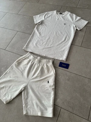 Vitt set från Polo Ralph Lauren - Snyggt vitt set från Polo Ralph Lauren med t-shirt och shorts. Båda plaggen har den klassiska lilla logotypen broderad på bröstet respektive benet. Tillverkade i mjukt bomullsmaterial för en bekväm känsla. Perfekt för en avslappnad och stilren look.