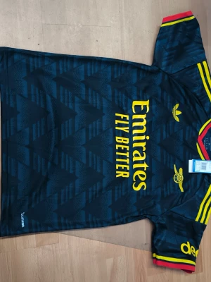 Arsenals nya bortaställ för säsongen 2026/2027 - Snygg Arsenal matchtröja i svart med gula detaljer och röda inslag på ärmarna. Adidas-logga och klubbmärke på bröstet, samt Emirates Fly Better-tryck. Nummer 14 och namnet Gyökeres på ryggen. Perfekt för fotbollsfans!