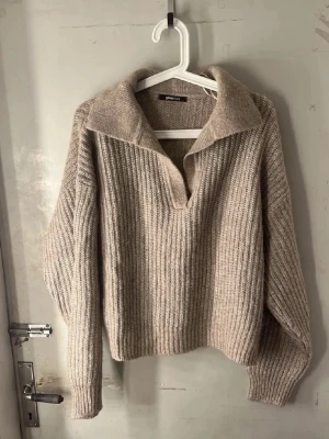 Beige stickad tröja med krage - Mysig beige stickad tröja med bred krage och djup v-ringning. Tröjan har ribbstickad struktur, långa ärmar och avslappnad passform. Perfekt för lager på lager och en chill look. Super mysig tröja att ha nu i höst!🤎