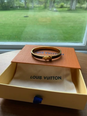 Louis Vuitton brunt armband i metall - Snyggt armband från Louis Vuitton i brunt och guldigt metallmaterial. Armbandet har ett klassiskt LV-mönster och ett dekorativt spänne framtill. Perfekt accessoar för dig som gillar exklusiv och tidlös design.