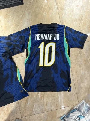 Brasilien Neymar Jr Bortaställ #10 - S - Snygg blå Brasilien matchtröja med Neymar Jr och nummer 10 på ryggen. Tröjan har gula och gröna detaljer, korta ärmar och officiellt landslagsemblem på bröstet. Tillverkad i lätt och ventilerande material, perfekt för fotboll eller träning.