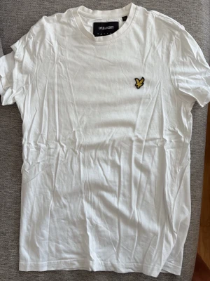 Vit t-shirt från Lyle & Scott - Klassisk vit t-shirt från Lyle & Scott med rund halsringning och den ikoniska gula örnloggan broderad på bröstet. Tillverkad i mjuk bomull för en skön känsla och enkel att matcha med allt. Perfekt basplagg för garderoben.