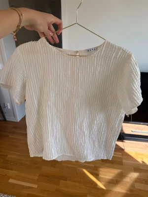 Vit strukturerad blus från Wera - Säljer en vit blus från Wera med korta ärmar och rund hals. Blusen har ett snyggt strukturerat tyg med diskreta ränder och en knappdetalj i nacken. Perfekt till jeans eller kjol för en clean och fräsch look.🤍