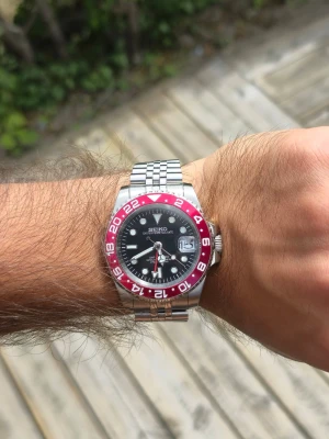 Seiko GMT red mod - Helt ny Seiko GMT Red mod som är väldigt stilren och sticker ut med sin helröda GMT-bezel 🔴✨  Boetten är 39 mm och ger en riktigt bra passform på handleden ⌚️ Svart urtavla tillsammans med den röda bezeln ger klockan en riktigt snygg look 🔥 Jubileelänk i rostfritt stål och datumvisning med förstoringsglas ✅ GMT-visare för extra tidszon 🌍  En riktigt snygg och unik klocka för dig som gillar Seiko mods ✨  Skickas välpackad 📦 Kontakta gärna för fler bilder eller mer info!