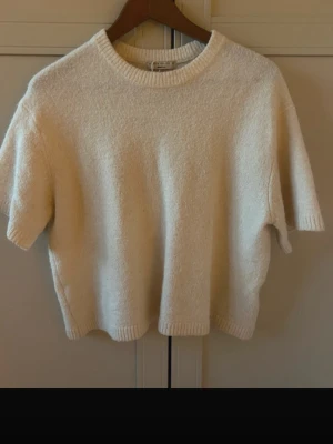 Krämvit stickad t-shirt - En krämvit stickad t-shirt med korta ärmar och rund hals. Tröjan har en avslappnad passform och ribbade kanter vid hals, ärmslut och nederkant. Perfekt för lager på lager och enkel att styla till olika looks.