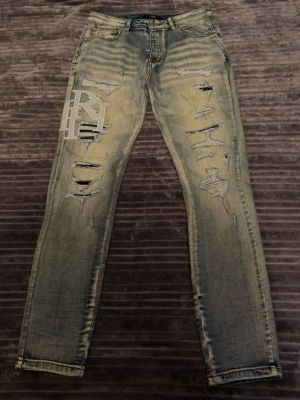 Distressed jeans från Amiri - Säljer ett par snygga ljusblå distressed jeans från Amiri med slitningar och patchade hål på båda benen. Jeansen har en rak passform och ett stort vitt tryck med bokstäver på vänster lår. Perfekta för dig som gillar streetwear och unik stil.