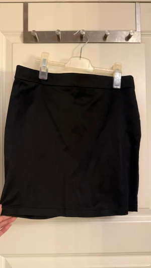 Svart kortkjol i enkel design - En stilren svart kortkjol med rak passform och slät midja. Kjolen har en minimalistisk look utan synliga detaljer eller mönster, vilket gör den lätt att matcha med olika stilar. Perfekt för dig som gillar en clean och klassisk vibe.