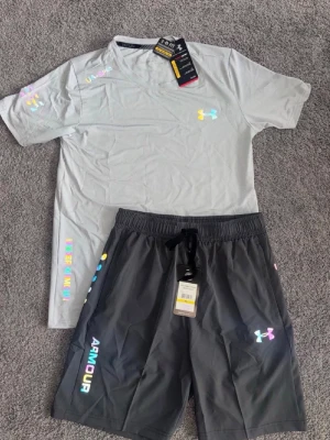 Grått träningsset från Under Armour - Snyggt träningsset från Under Armour med ljusgrå t-shirt och mörkgrå shorts. Setet har färgglada loggor och tryckdetaljer på både tröja och shorts. T-shirten har rund hals och korta ärmar, shortsen har resår i midjan och dragsko. Perfekt för gym eller löpning.