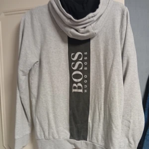 Grå hoodie från Hugo Boss - Snygg grå hoodie från Hugo Boss med dragkedja framtill och svart logga på bröstet. Stora BOSS-trycket på ryggen ger en cool look. Huva med svarta snören och två fickor. Tillverkad i mjukt bomullsmaterial för skön känsla.