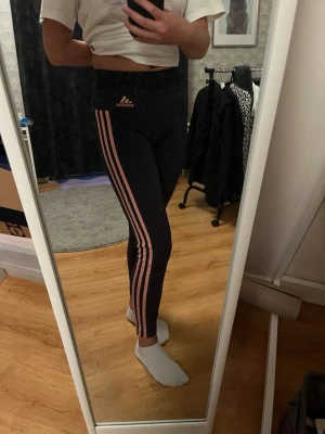 Svarta Adidas leggings med rosa ränder - Säljer ett par svarta Adidas leggings i bomull med egt brett midjeband och tre rosa ränder längs benen. Adidas-logga i rosa framtill. 