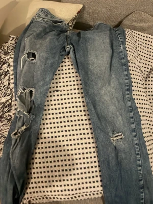 Blå slitna jeans med hål - Säljer ett par blå jeans med flera stora hål och slitningar på benen. Jeansen har klassisk femficksmodell, normal midja och dragkedja med knapp. Perfekta för en avslappnad och trendig look.