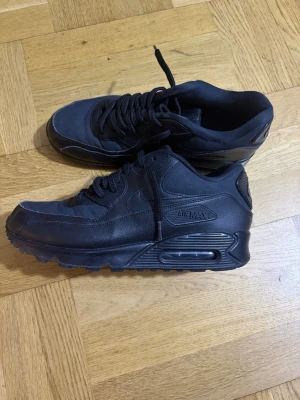 Svarta Nike Air Max 90 sneakers - Säljer ett par helsvarta Nike Air Max 90 sneakers med klassisk chunky sula och synlig Air-enhet. Skorna är tillverkade i en mix av läder och textil, har svarta skosnören och diskreta Nike-loggor på sidan. Perfekta för dig som gillar en clean och sportig look.