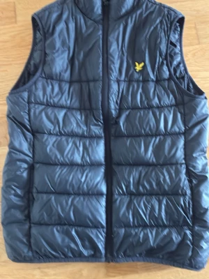 Svart väst från Lyle & Scott - Snygg svart väst från Lyle & Scott med gul logga på bröstet. Västen har hög krage, dragkedja framtill och quiltad design. Värmer men är ändå ganska tunn. Passar ovanpå en tröja, tunnare jacka eller som en värmande väst under jackan. Sparsamt använd. Hittar inga fel eller synliga skador men använd