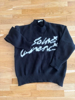 Svart stickad tröja Saint Laurent - Svart stickad tröja från Saint Laurent med vit text i grafisk stil över bröstet. Tröjan har ribbstickad krage, ärmslut och nederkant. Mjuk och lite oversized passform, perfekt för en trendig look.