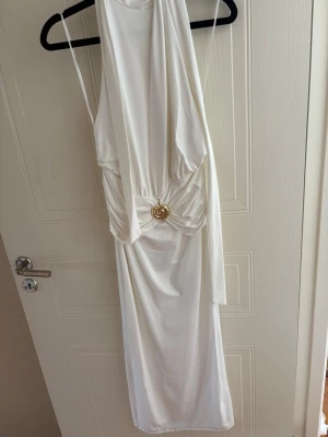 Vit långklänning med gulddetalj - Elegant vit långklänning i mjukt, glansigt material. Klänningen har ärmlös design, draperad midja och en dekorativ guldfärgad detalj framtill. Perfekt för speciella tillfällen när du vill sticka ut.
