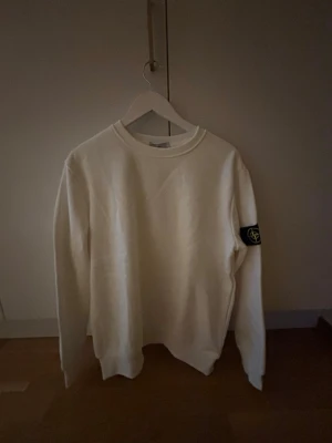 Stone island vit sweatshirt - Vit hoodie med rund hals och ribbade muddar. Klassisk och stilren design med en svart patch med logga på vänster ärm. Perfekt för en avslappnad och trendig look. Passar bättre på L