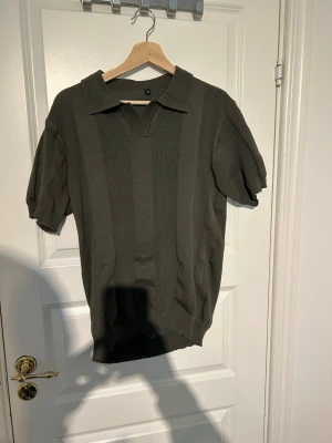 Olivgrön stickad pikétröja - Snygg olivgrön stickad pikétröja med korta ärmar och klassisk krage. Tröjan har ett diskret ribbat mönster i vertikala paneler och är tillverkad i ett mjukt, bekvämt material. Perfekt för en stilren och avslappnad look.