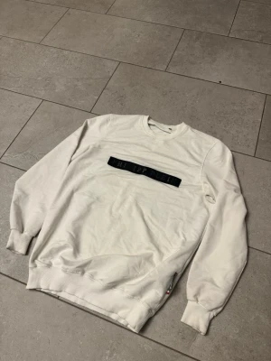 Vit sweatshirt från Philipp Plein - Snygg vit sweatshirt från Philipp Plein med svart logotypband över bröstet. Tröjan har rund halsringning, långa ärmar och ribbade muddar vid ärmslut och nederkant. Perfekt för en clean och stilren look.