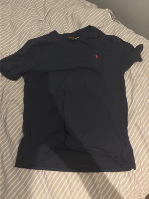 Mörkblå t-shirt från Polo Ralph Lauren - Säljer en mörkblå t-shirt från Polo Ralph Lauren med klassisk röd logga broderad på bröstet. T-shirten har rund hals och korta ärmar, perfekt för en clean och enkel stil. Tillverkad i mjuk bomull för skön känsla hela dagen.
