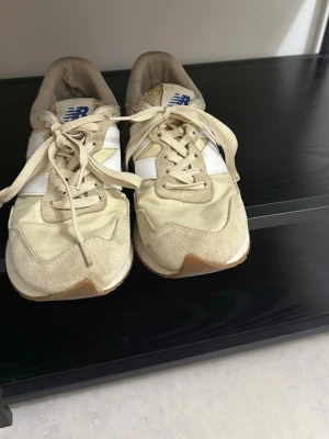 Beige New Balance sneakers - Snygga beige sneakers från New Balance med vita detaljer och klassisk logga på plösen. Skorna har låga skaft, snörning och en mix av mocka och textilmaterial. Perfekta för en avslappnad och trendig stil.
