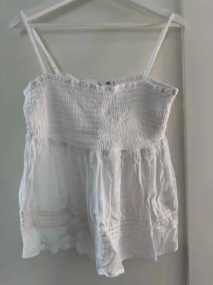 Vit linnetopp med spetsdetaljer - Söt vit linnetopp med smala axelband och smockad överdel. Toppen har volangkant nedtill och fina spetsdetaljer längs fållen. Perfekt för varma dagar och enkel att styla med jeans eller kjol. Storlek M från lager 157. Aldrig kommit till användning tyvärr, därav nyskick! 