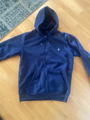 Mörkblå hoodie från Polo Ralph Lauren - Mörkblå hoodie med dragkedja från Polo Ralph Lauren. Klassisk design med vit broderad logga på bröstet, två stora fickor framtill och justerbar huva. Tillverkad i mjukt bomullsmaterial som känns skönt mot huden. Perfekt för en avslappnad och stilren look.