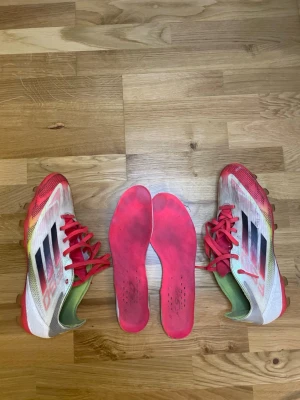 Vita och röda Adidas fotbollsskor f50 pro - Säljer ett par Adidas fotbollsskor i vitt och rött med svarta detaljer och röda snören. Skorna har en låg profil och är designade för spel på gräs. Yttermaterialet är syntetiskt och sulan har dobbar för bra grepp. Innersulorna är löstagbara och röda.