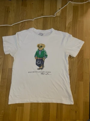 Vit Polo Bear t-shirt från Ralph Lauren - Vit t-shirt från Ralph Lauren med ikoniskt Polo Bear-tryck framtill. Björnen har solglasögon, grön jacka och randig tröja. Klassisk rund hals och korta ärmar. Perfekt för dig som gillar streetstyle och vill ha något unikt i garderoben. storlek 160-170 