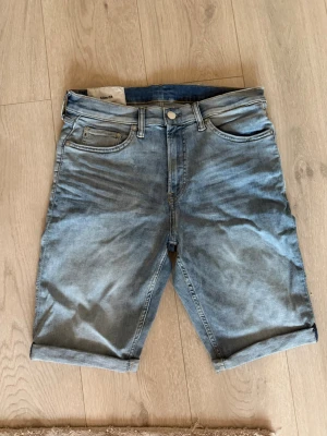 Blå denimshorts med uppvik - Säljer ett par klassiska blå denimshorts med fem fickor och uppvikta benslut. Shortsen har normal passform och stängs med knapp och dragkedja. Perfekta för varma dagar och enkla att matcha med olika stilar.