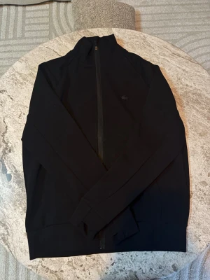 Lacoste Kofta Zip - Snygg svart Lacoste-kofta med dragkedja framtill och klassisk krage. Koftan har en stilren design med den ikoniska Lacoste-krokodilen på bröstet. Perfekt för dig som vill ha en exklusiv och stilren look i garderoben. Den är liten i storleken så den passar XS, Även ifall det står S. Helt ny