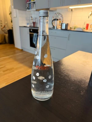 Evian x Moncler vattenflaska glas - Snygg glasflaska från Evian i samarbete med Moncler. Flaskan är genomskinlig med vita prickar och har skruvkork i metall. Perfekt som samlarobjekt eller dekoration i köket.