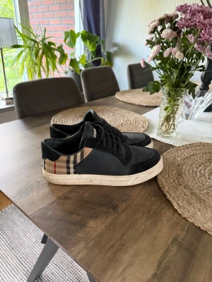 Svarta Burberry sneakers med rutigt mönster - Snygga svarta sneakers från Burberry med klassiskt beige, svart och rött rutmönster på sidan. Skorna har svarta snören, vit sula och är tillverkade i en mix av mocka och textil. Perfekta för dig som vill ha en stilren och trendig look.