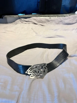 Svart bälte med silverfärgat vargspänne - Snyggt svart bälte i skinn med ett unikt silverfärgat spänne i form av ett varghuvud. Perfekt accessoar för att ge din outfit en cool och edgy touch. Bältet har klassisk form och flera hål för justerbar passform.