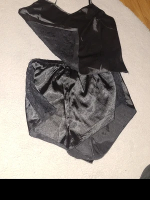 Svart satinpyjamas set med spets - Snyggt svart pyjamasset i satin med linne och shorts. Linnet har tunna axelband och shorts med elastisk midja. Båda delarna har fina spetsdetaljer på kanterna för en extra elegant look. Perfekt för en mysig kväll hemma.