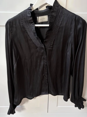 Neo noir blus - Stilren svart blus från neo noir med långärmad design, volangdetaljer vid kragen och smockade muddar vid ärmsluten. Blusen har knäppning framtill och är gjord i ett glansigt, mjukt material som ger en lyxig känsla.