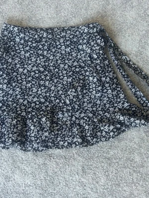 Svart blommig omlottkjol med volang - Söt svart omlottkjol med vita små blommor och volangkant. Kjolen har knytband i sidan och är perfekt för dig som gillar en feminin och trendig stil. Lätt och luftig, passar till många olika outfits.