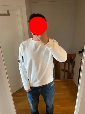 CP company sweatshirt - Säljer en stilren vit sweatshirt med rund hals och ribbade muddar. Tröjan har en unik detalj med en dragkedjeförsedd ficka på vänster ärm. Perfekt för dig som gillar enkel men snygg design.