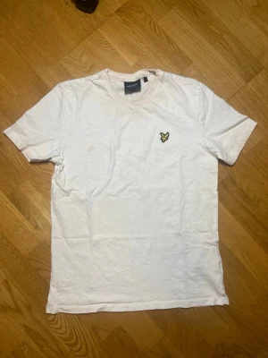 Vit t-shirt Lyle & Scott - Säljer denna klassiska vita t-shirt från Lyle & Scott med rund halsringning och den ikoniska gula örnloggan broderad på bröstet. Tillverkad i mjuk bomull och har en enkel, stilren design som passar till det mesta. OBS! Det finns en märkbar matfläck på bröstet och lite röd missfärgning runt halsringningen. Jag har kollat upp det och det går att få bort det med en kemisk blandning och inte med traditionell tvättmaskin. Vid frågor, extra bilder eller liknande är det bara att kontakta mig!