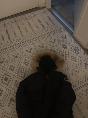 Svart dunjacka med patch - Snygg svart canda zavetti dunjacka med quiltad design och stor patch på ärmen där det står 'Zavet Canada'. Jackan har hög krage, dragkedja och knappar framtill samt två stora fickor. Perfekt för kalla vinterdagar. L junior storlek 