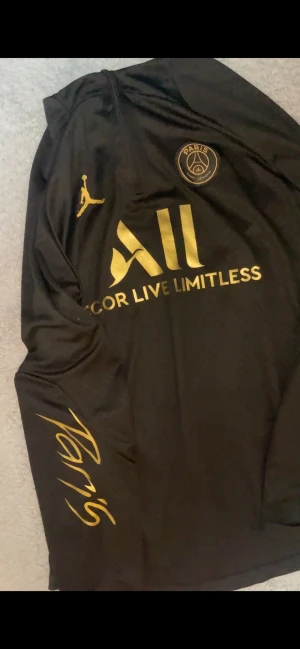 Svart PSG långärmad träningströja - Snygg svart långärmad träningströja med guldiga detaljer från Paris Saint-Germain. Tröjan har PSG-logga, Jordan-logga och texten 'Accor Live Limitless' samt 'Paris' på ärmen. Tillverkad i ett lätt och sportigt material som passar perfekt till träning.
