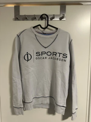 Grå sweatshirt Oscar Jacobson - Grå sweatshirt från Oscar Jacobson med svart SPORTS-tryck på bröstet. Tröjan har rund halsringning, ribbade muddar och dekorativa sömmar i kontrastfärg. Perfekt för en sportig och avslappnad stil.