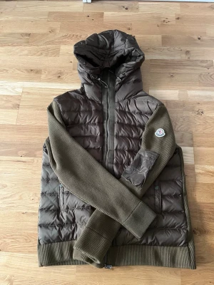Cardigan - Snygg brun/grön cardigan med stickade, olivgröna ärmar och ribbade muddar. Jackan har en vadderad kropp, dragkedja framtill, två sidofickor och en huva. Storleken står att det är XL inuti men passar för mindre som t.ex M. 
