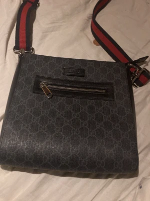Svart axelväska med GG-mönster - Snygg svart axelväska från gg med klassiskt GG-mönster över hela väskan. Väskan har en framficka med dragkedja och en bred axelrem i rött och marinblått. Materialet är tåligt och har en lyxig känsla. Perfekt för dig som vill ha en stilren och trendig väska.